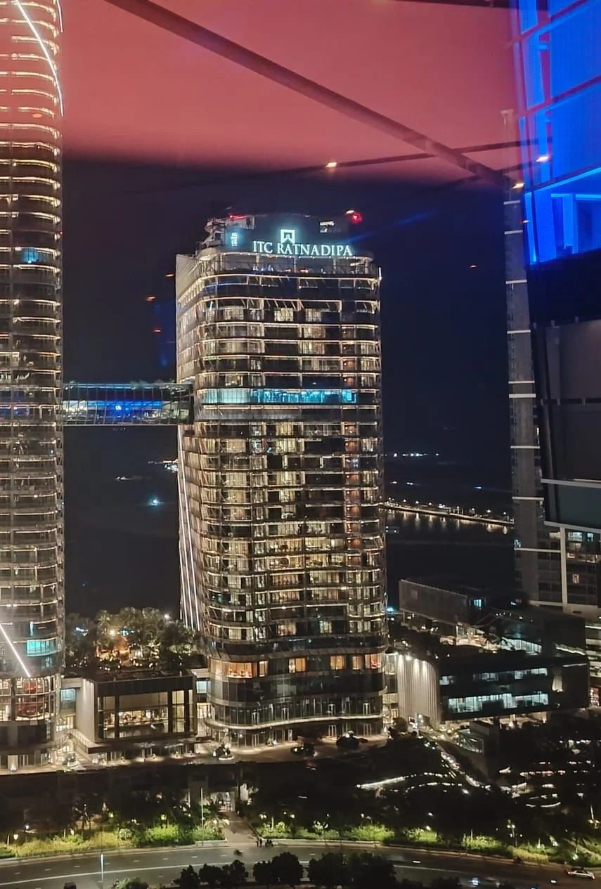 Night view of a cityscape with illuminated futuristic high-rise buildings, including the ITC Rajputana hotel at the top of the central building, and a bridge over water in the background.