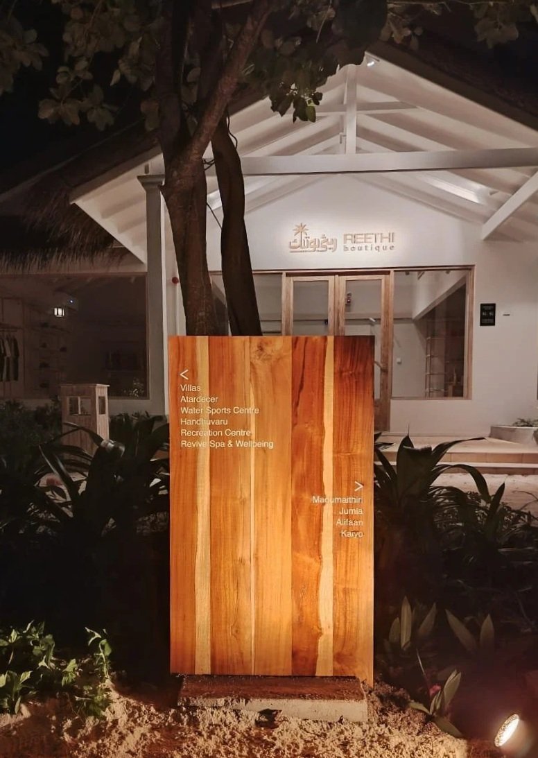 A wooden signboard displaying location information in front of a white building labeled 'Reethi Boutique' at night. The sign lists various amenities and nearby attractions, and is surrounded by plants and lit from below.