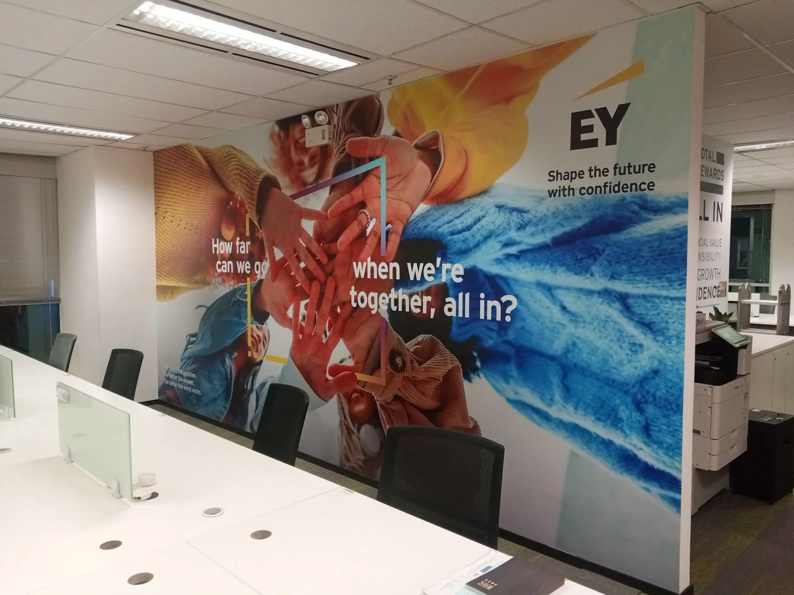 Office conference room with a large EY poster on the wall showing multiple hands joined together, a person smiling, and a wave. The poster has text: "How far can we go when we're together, all in?" and EY logo with the tagline "Shape the future with confidence."