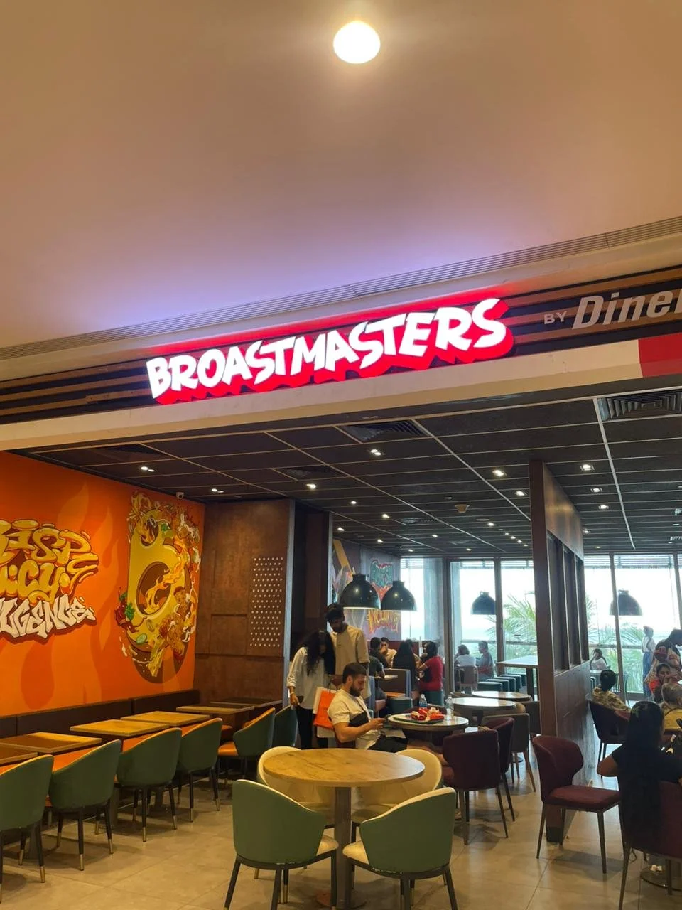 Interior view of a restaurant called Broastmasters, showing seating arrangement, customers dining, and colorful wall art with a pizza graphic.