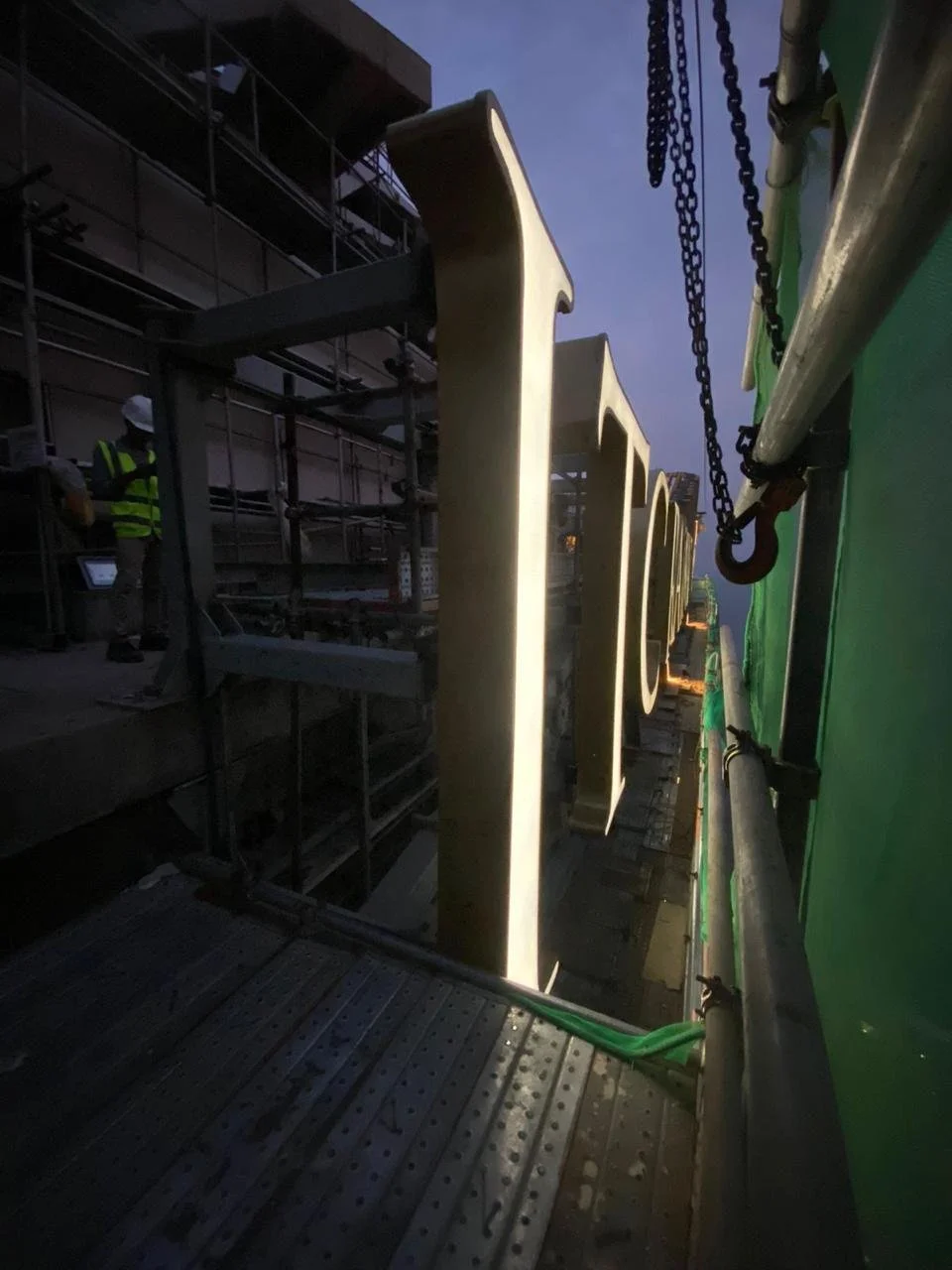 Close-up of large illuminated outdoor signage with metal framework and green protective covering, at a construction site during dusk, with construction workers in safety vests and helmets in the background.