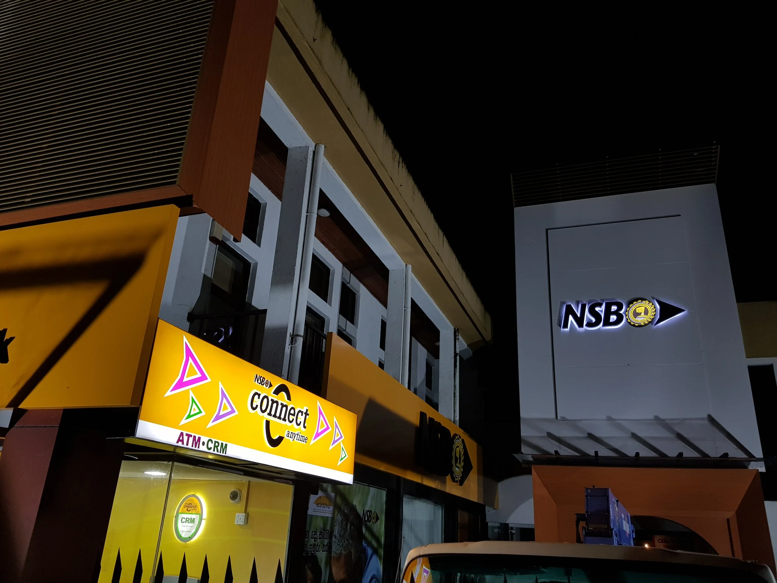 Nighttime view of storefront with illuminated yellow and green signs for 'Connect' and 'CRM', part of a business complex, with building exterior and signage visible.