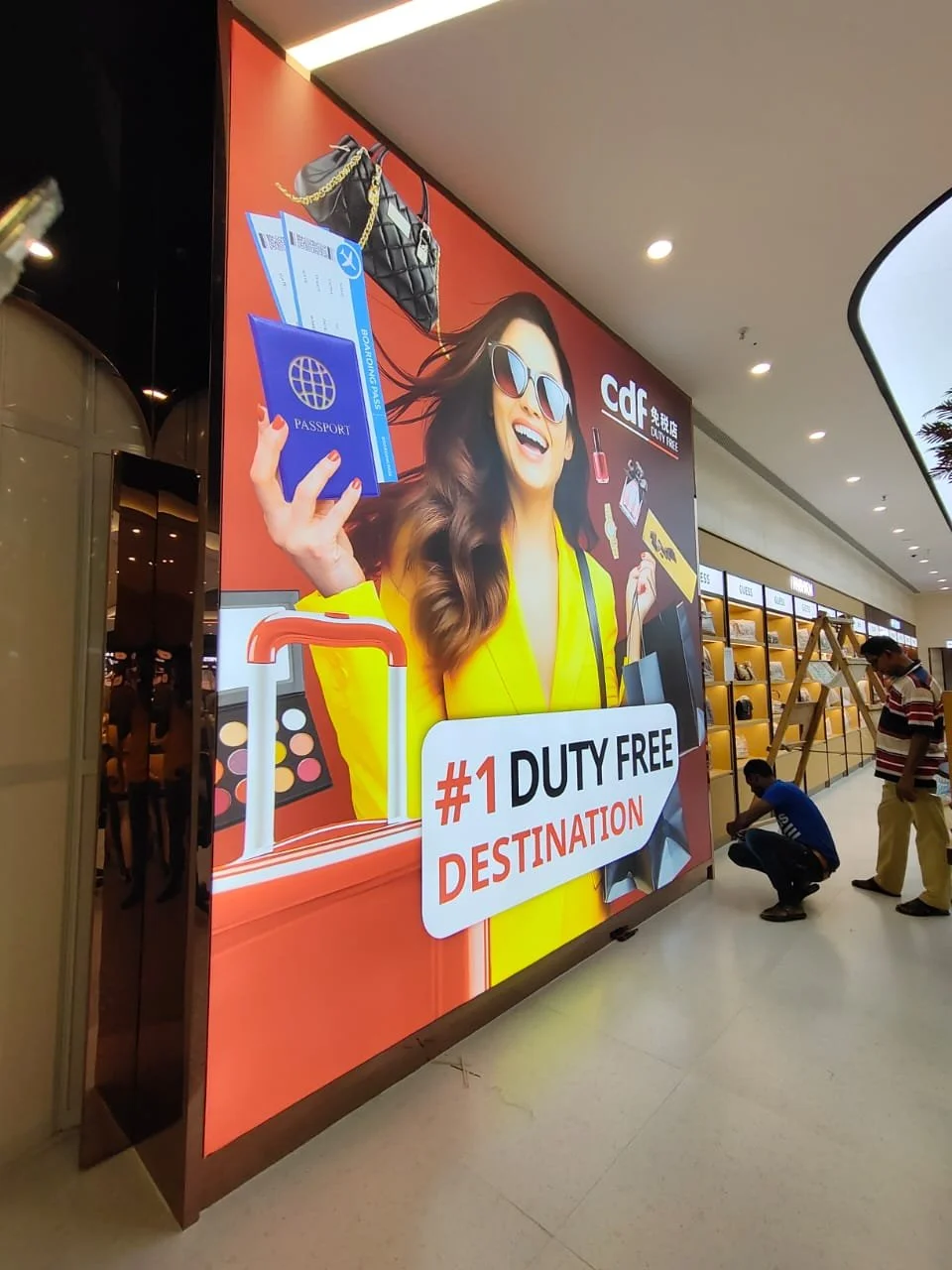 Large advertising sign promoting duty-free shopping at an airport, featuring a woman in sunglasses holding a passport and boarding pass. The sign includes images of a handbag, makeup, and travel essentials, with text '#1 Duty Free Destination.'