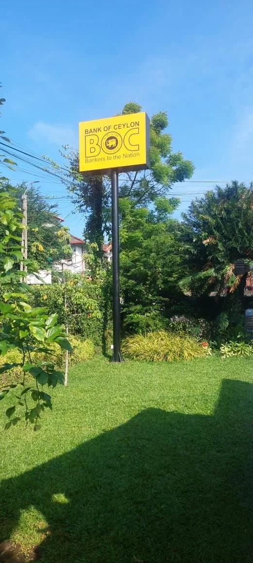 Sign for Bank of Ceylon (BOC) with a yellow background and black text, located outdoors with greenery and blue sky.