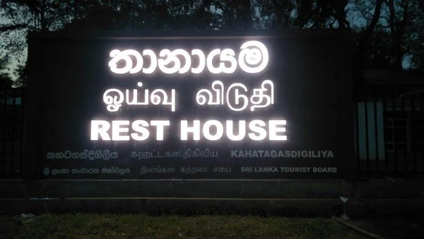 Signboard with the word 'Rest House' in English and similar scripts in a tropical outdoor setting.