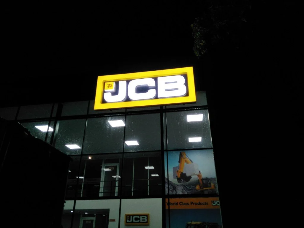 Nighttime exterior view of a JCB store with a large illuminated sign on the building showing the JCB logo in white letters within a yellow rectangle. The store has glass windows showing indoor lighting and construction equipment images.