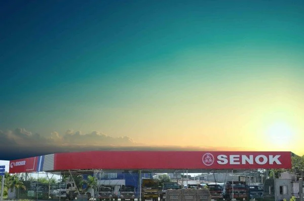 A large red and white canopy with the word 'SENOK' and a logo, set against a sunset sky with a parking area underneath.