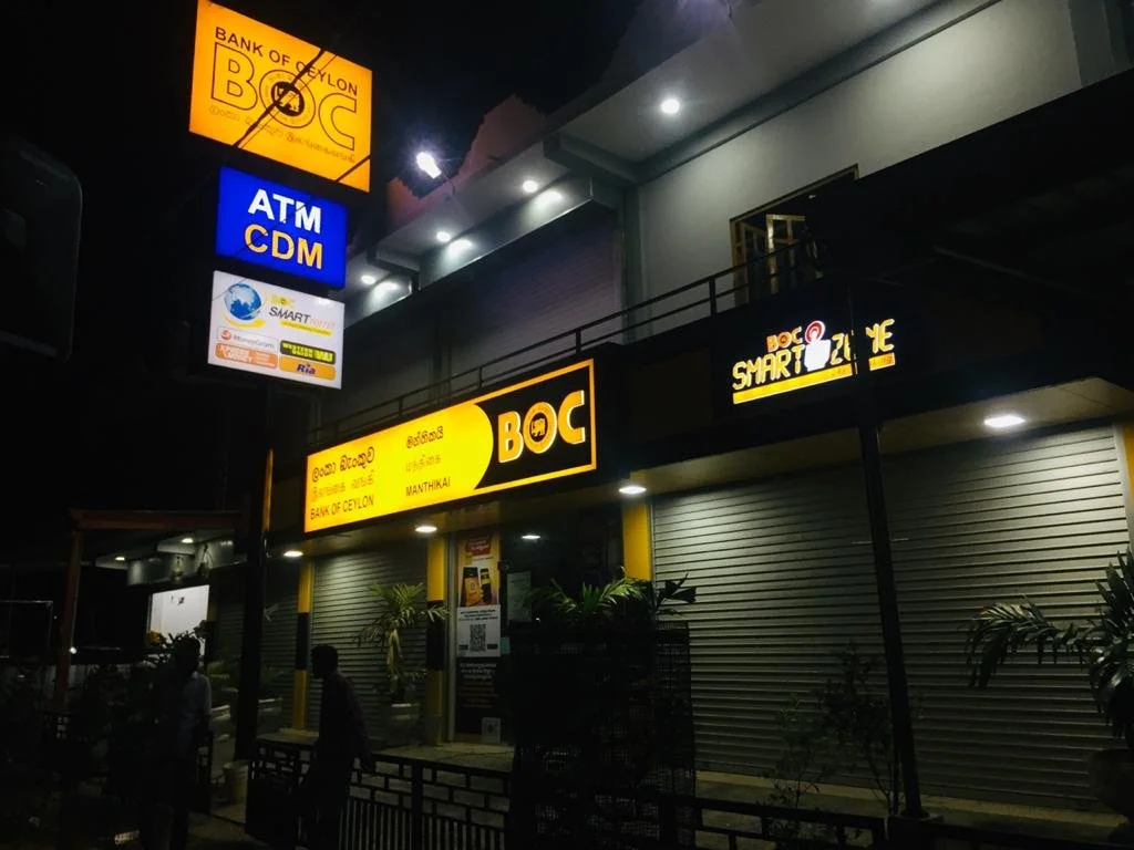 Nighttime scene of a closed bank and ATM with illuminated signs for Bank of Ceylon and a Smart customer service center, and a sign for BOQ Happy, with some plants and people walking by.