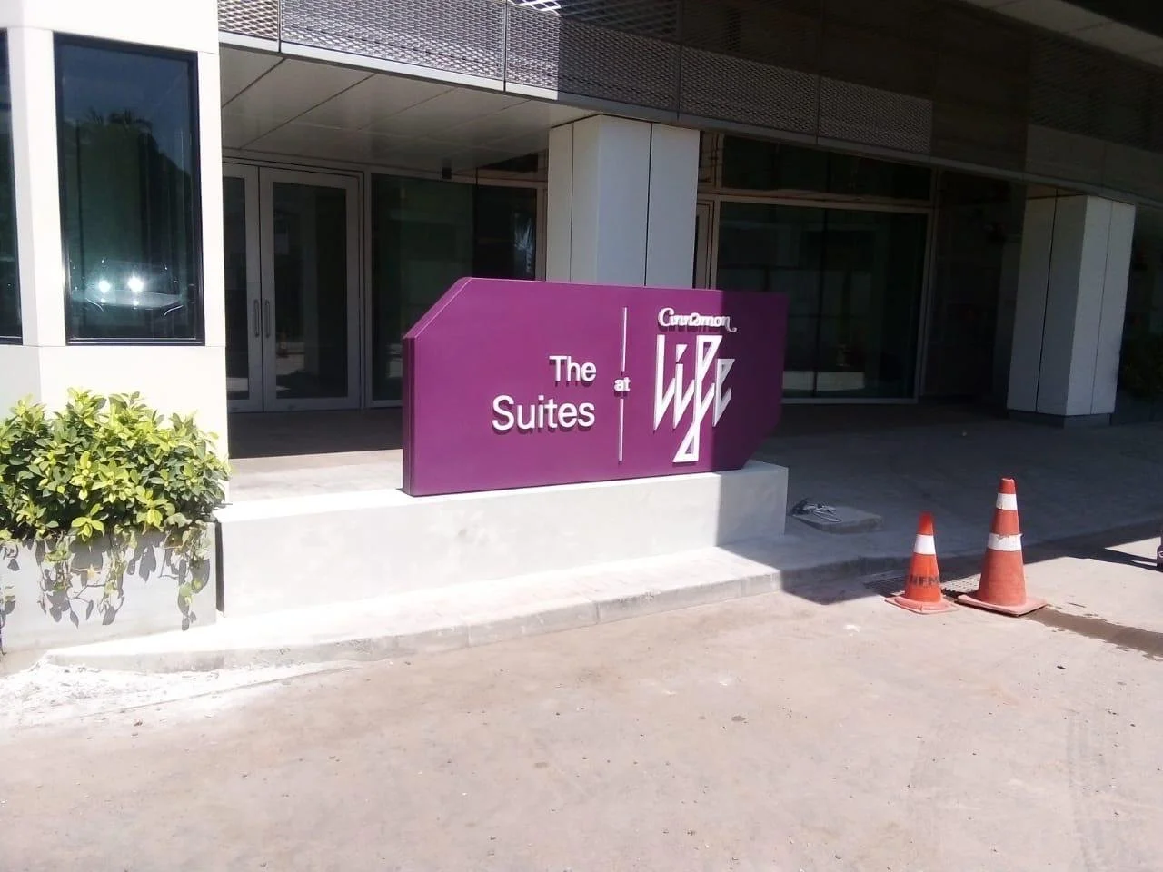 A purple sign reading 'The Suites at Cusamoor' outside a modern building under construction, with orange traffic cones on the sidewalk.