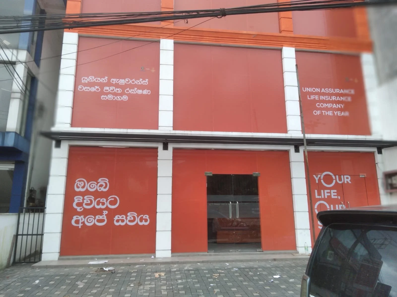The exterior of a building with red panels displaying text in Sinhala and English, including phrases like 'Union Assurance Life Insurance Company of the Year' and 'Your Life, Our Life'.