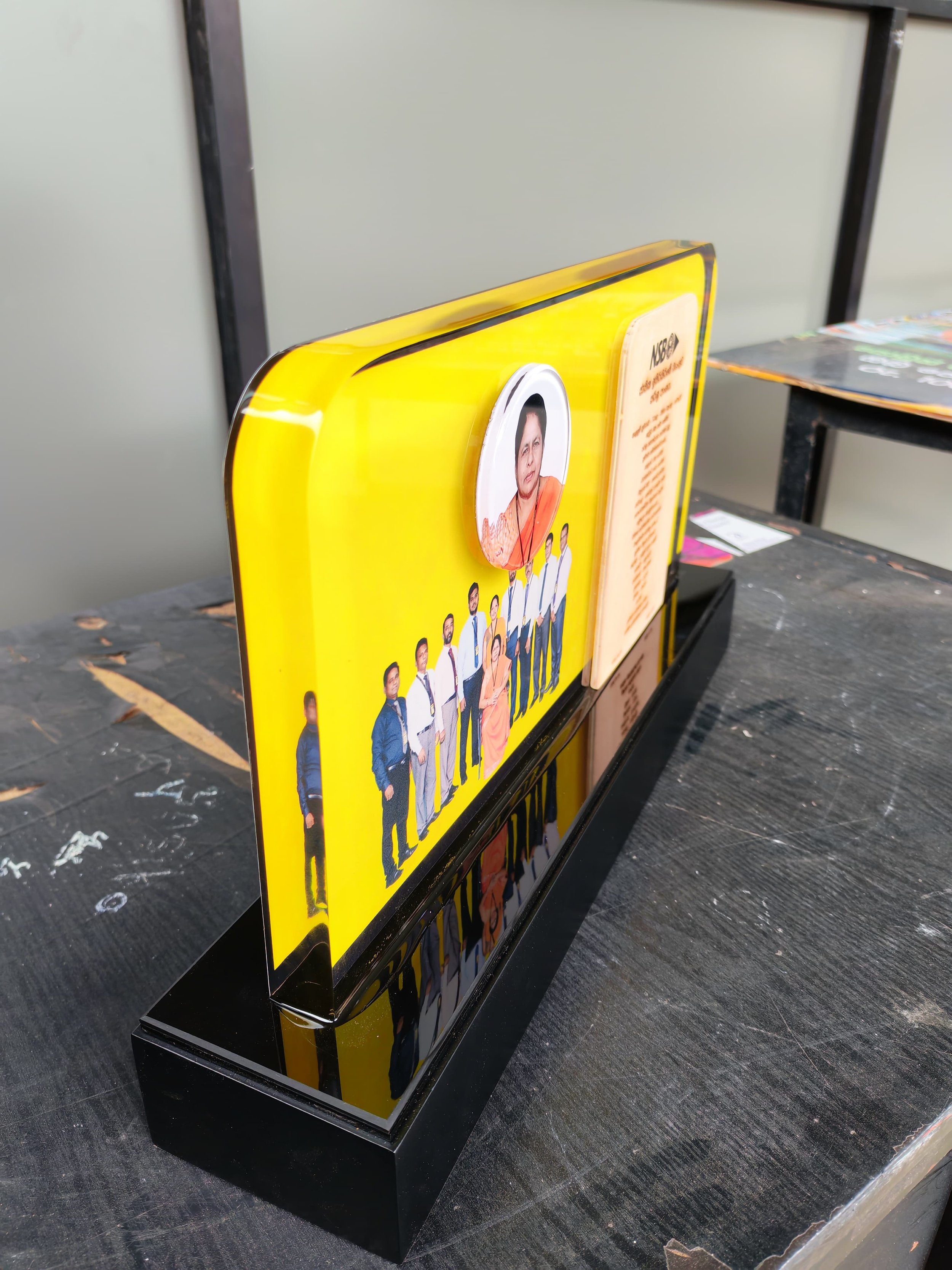 A yellow stand or display with a picture of an Indian woman in traditional attire and a group of young people dressed in light blue uniforms, on a black wooden surface, next to other magazines or brochures.