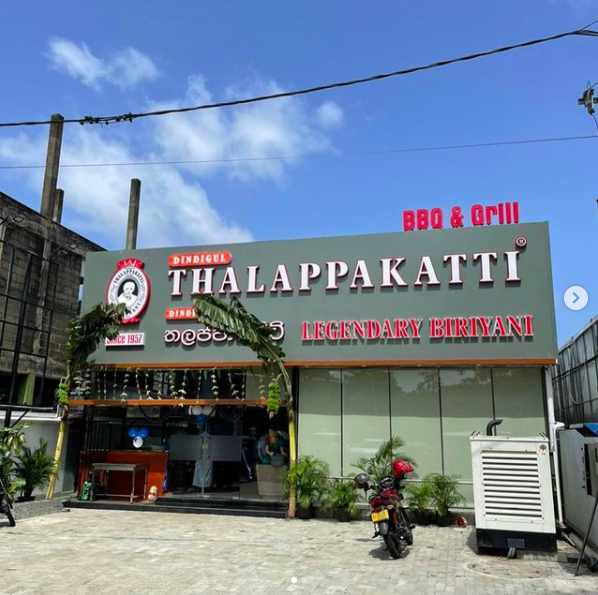 Signboard for Thalappakatti restaurant advertising BBQ and grill, legendary biryani, with outdoor seating and a motorbike parked outside.