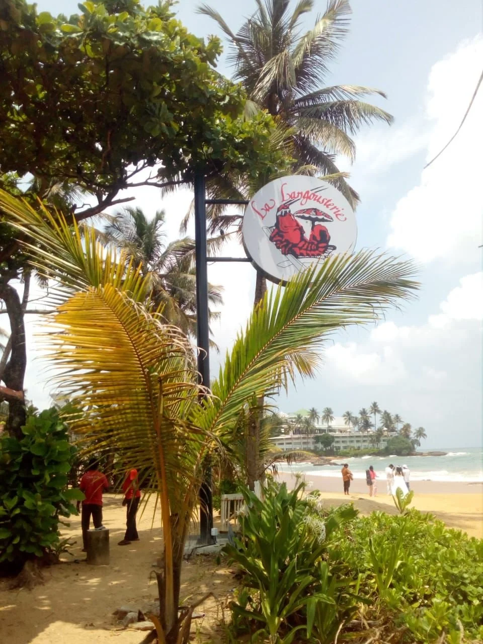 Beachside scene with palm trees, a sign for 'La Sangoustine', and people walking along the sandy beach with ocean waves in the background.