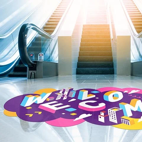 Escalator with a colorful floor decal of the word 'Welcome' in large, playful letters in an airport or shopping mall.