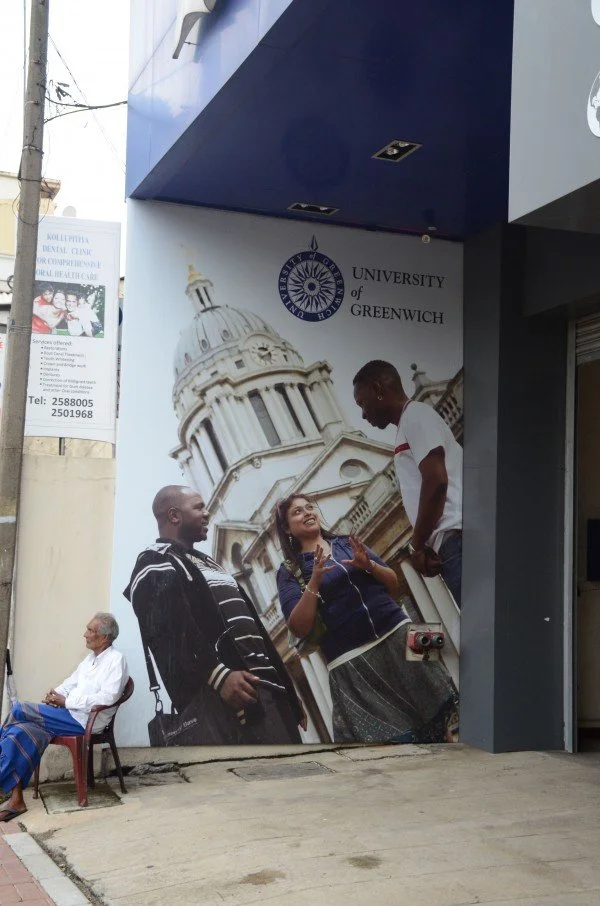 A large poster on an exterior wall shows three people, two men and one woman, standing and talking in front of a historical building with a dome. Above them is the logo for the University of Greenwich. Nearby, an older man with glasses is seated on a