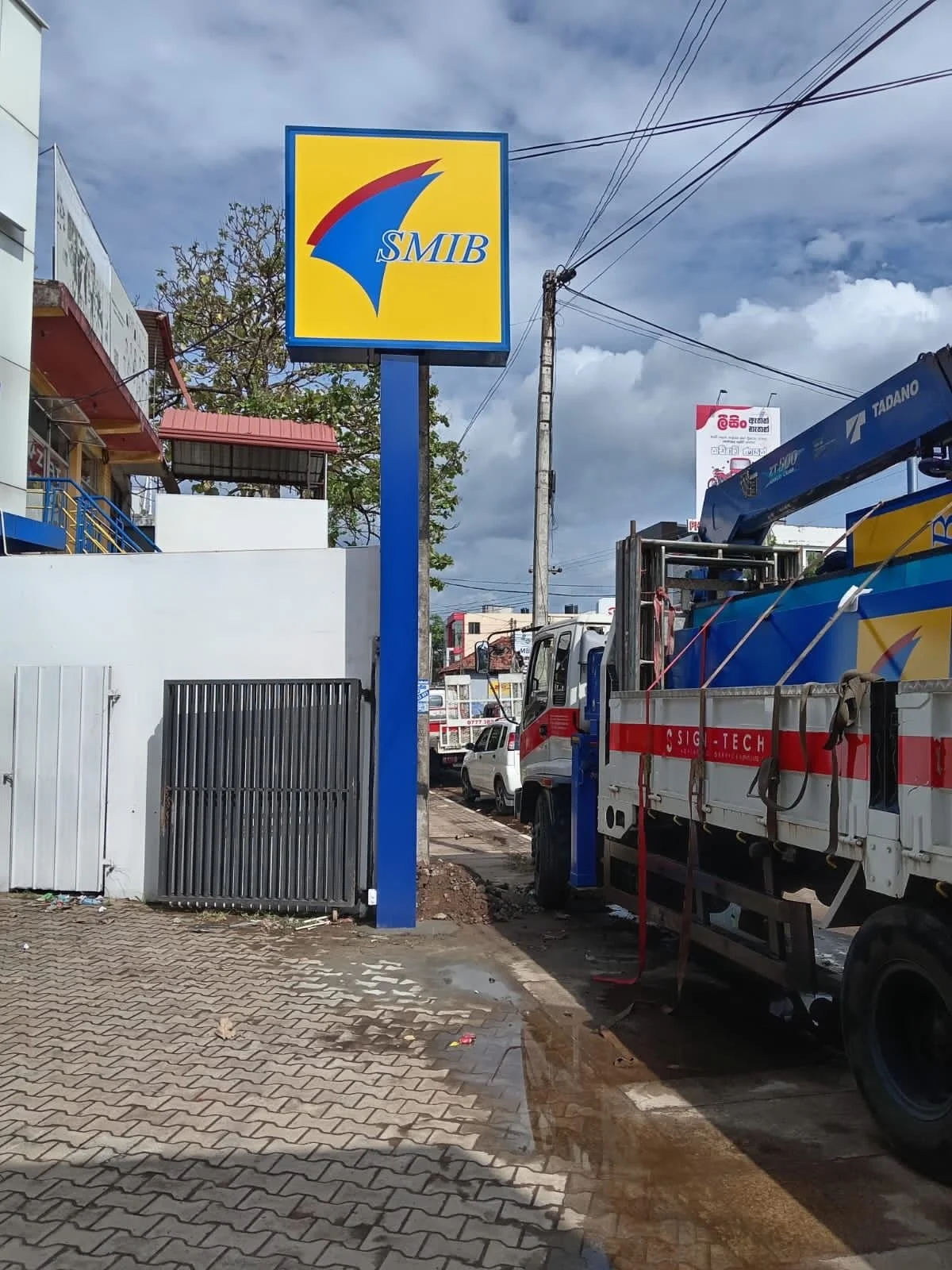 A street scene with a blue and yellow SMIB sign, construction work with trucks, and a cloudy sky.