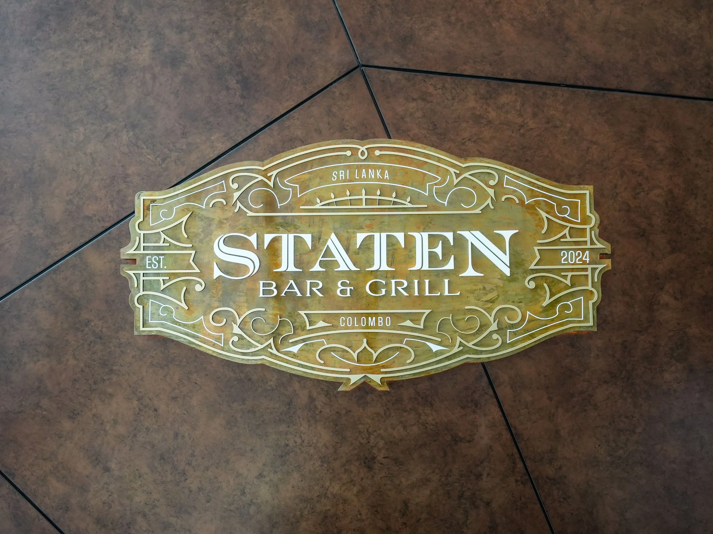 A decorative sign for Staten Bar & Grill in Colombo, Sri Lanka, with ornate gold and white design, on a brown tiled floor.