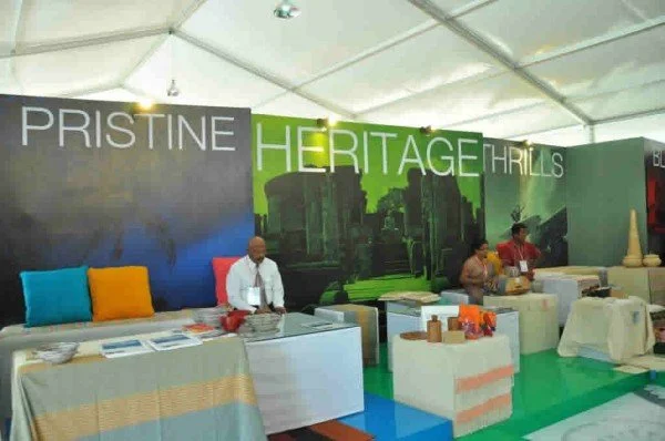 An exhibition booth with banners reading "Pristine Heritage Trails," featuring a table with promotional materials, and three people attending to the booth in a large tent.