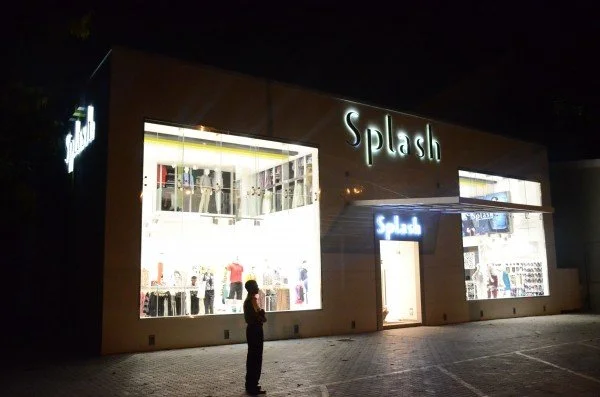 Nighttime view of a retail clothing store named Splash with large illuminated windows displaying mannequins and clothing inside, and a person standing outside.