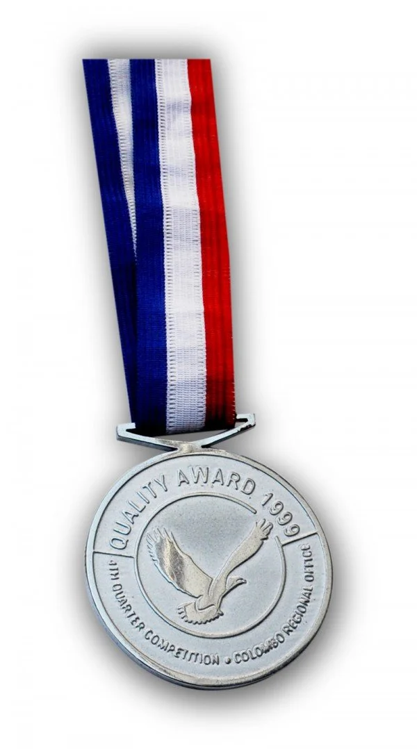A silver medal with a ribbon in red, white, and blue stripes. The medal is inscribed with "QUALITY AWARD 1999" and features an eagle in flight.