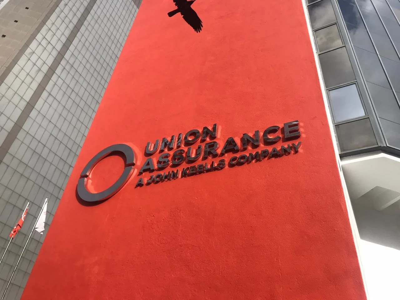 A large orange building wall featuring a black 3D logo of a circle and eagle, with black 3D text reading 'Union Assurance A John Keells Company' on the wall.