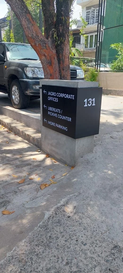 A black directional sign on a concrete base guiding to Jagro Corporate Offices, Ubereats/PickMe counter, and more parking, situated in an outdoor parking lot next to a tree and a parked vehicle.