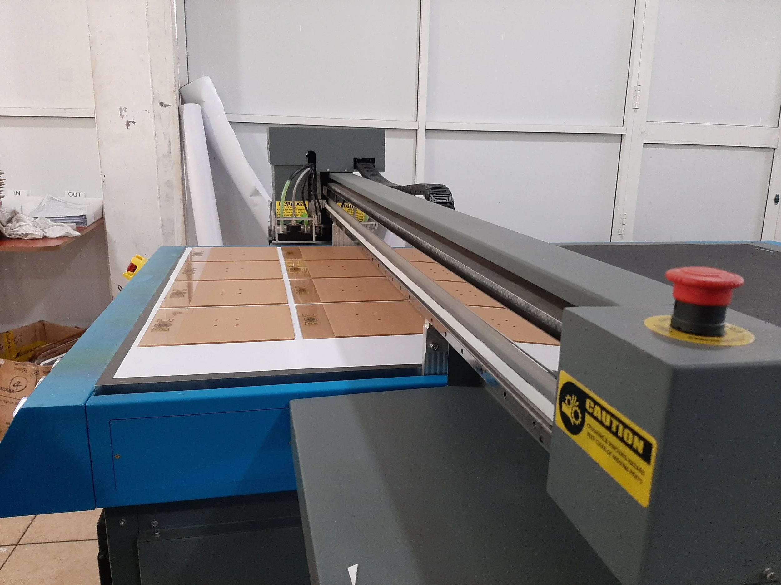 Industrial machine with a long metal arm in a workshop setting, yellow caution labels, and boxes with papers nearby.