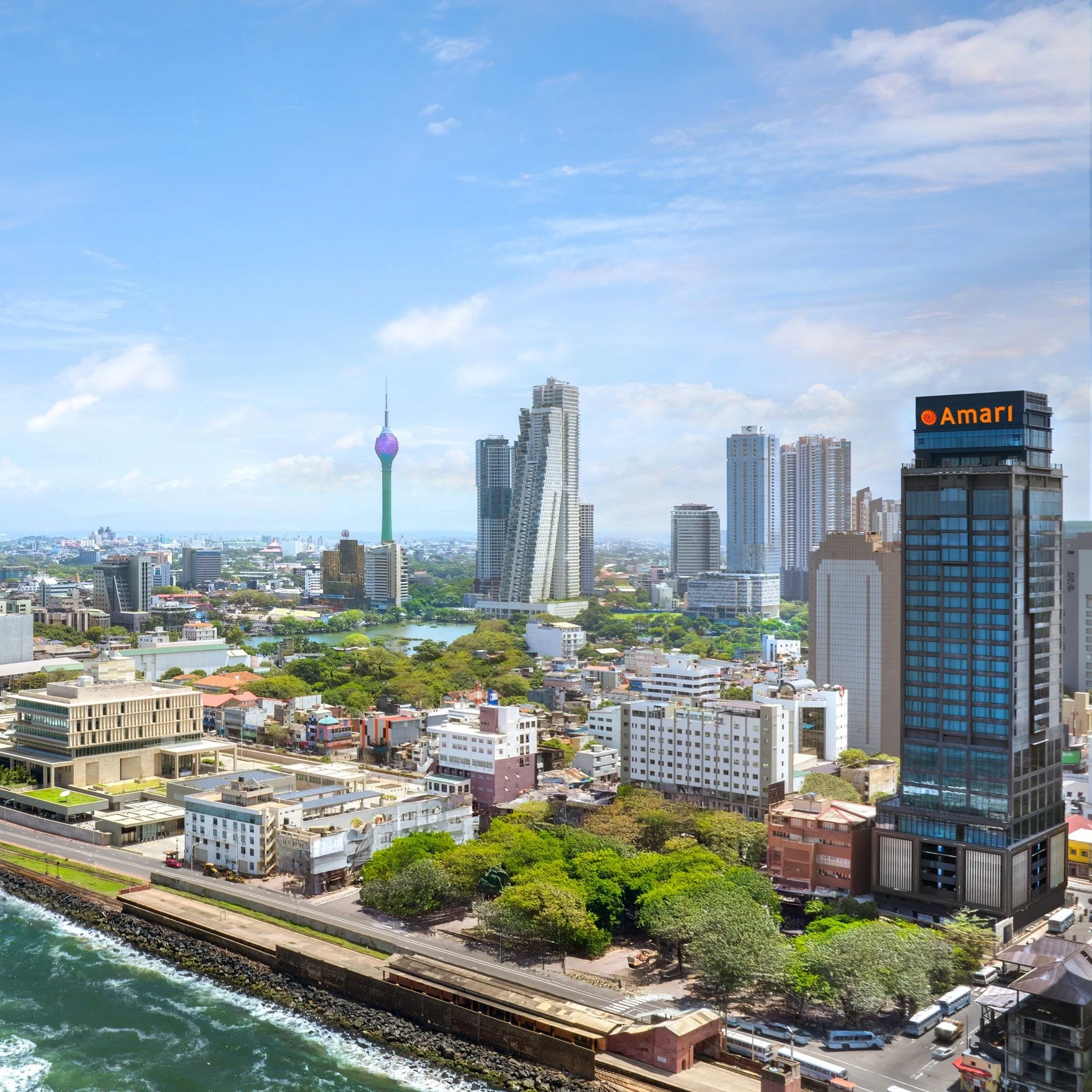 City skyline with tall modern buildings, a prominent tower with a colorful, spherical top, and a waterfront with a railway track and greenery.