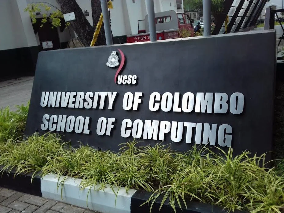 A black rectangular sign for the University of Colombo School of Computing with the university's logo at the top, placed on a small garden bed with green plants in front of it. Background includes trees, a street, and parked vehicles.
