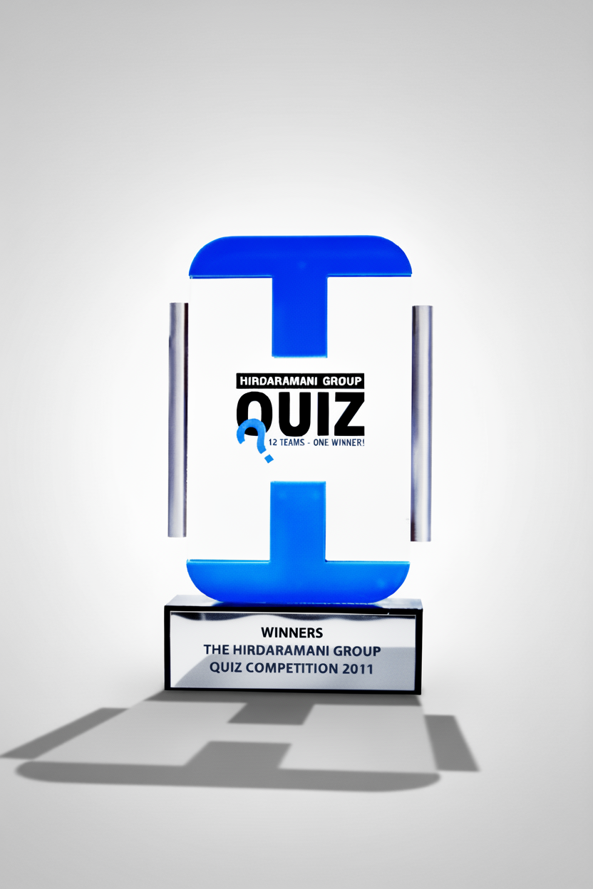 A trophy with a blue stylized letter 'T' on top, a black sign with 'Hirdaramani Group Quiz 12 Teams - One Winner!', and a black base with an inscription for the winners of the 2011 quiz competition.