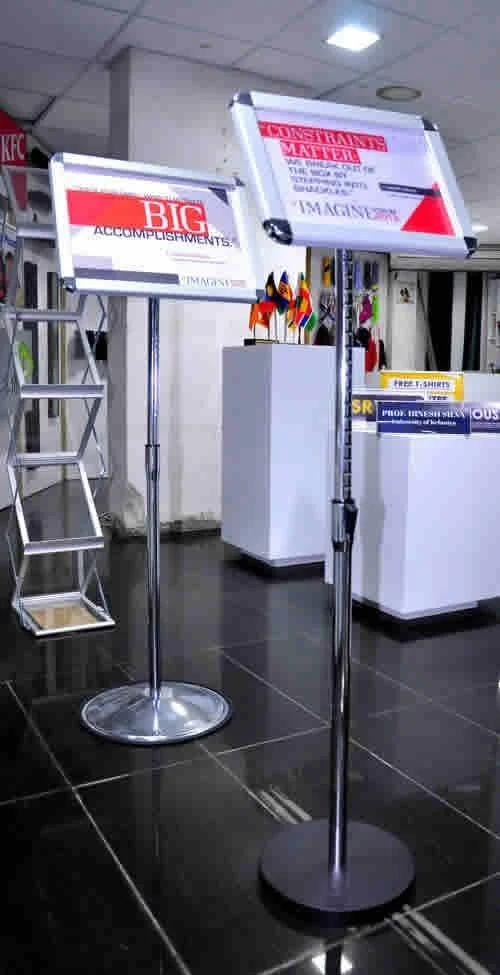 Two kiosks or stands with signs on top in a store or office setting, surrounded by dark tile flooring and white walls, with boxes and a colorful cat sculpture in the background.