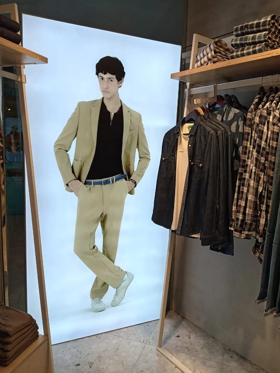 Clothing store display with a large poster of a man in a beige suit, black shirt, and white sneakers, surrounded by racks of plaid and denim shirts.