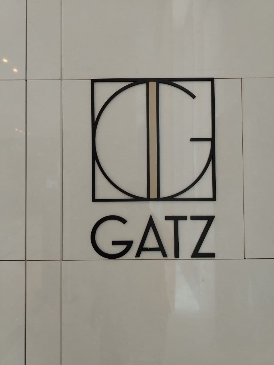 A modern wall sign with a stylized logo featuring a black square with a circle and line inside, and the word 'GATZ' below in black capital letters, mounted on a white tiled wall.