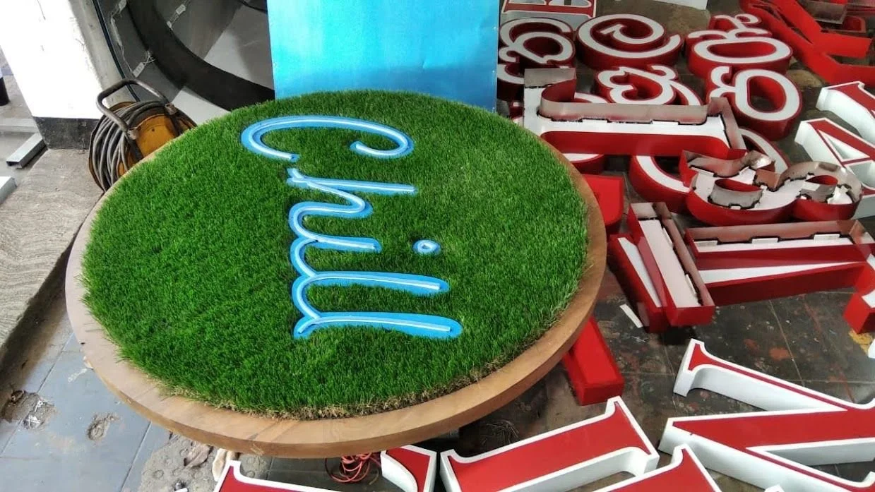 A round sign with a green grass background and blue neon text that says 'Chill'. Surrounding the sign are red and white letters that appear to be part of a larger display or signage project.