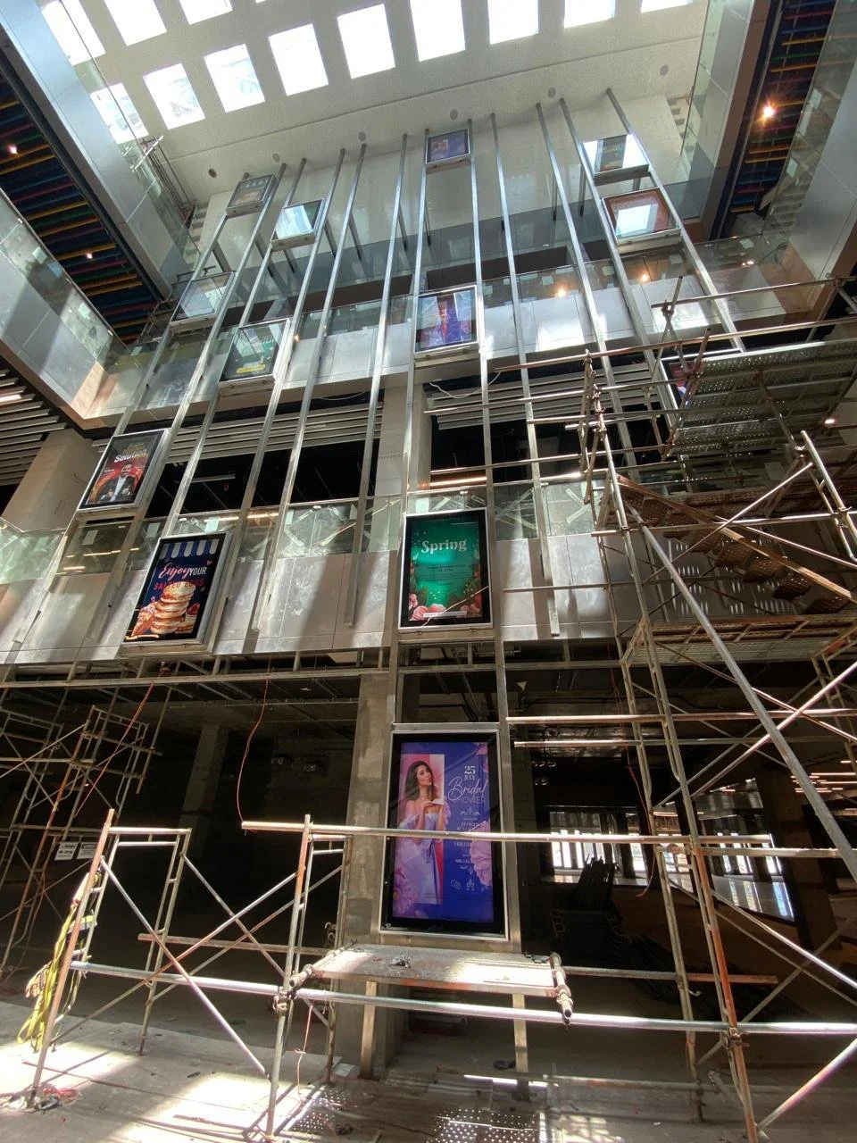 Interior of a building under construction with digital advertisement screens, scaffolding, and metal framework.