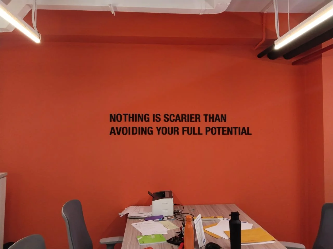 An orange wall with a motivational quote in black text reading, "Nothing is scarier than avoiding your full potential." A cluttered conference table with papers, folders, bottles, and office supplies, and gray chairs on either side.
