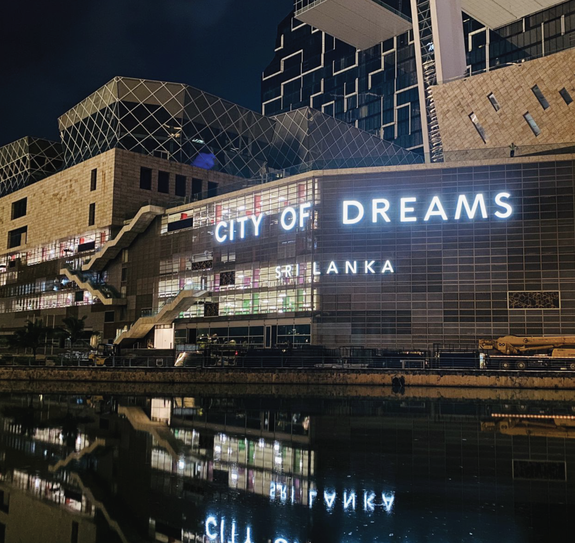 Night view of a modern multi-story building with illuminated sign saying 'City of Dreams', reflecting on a water surface.