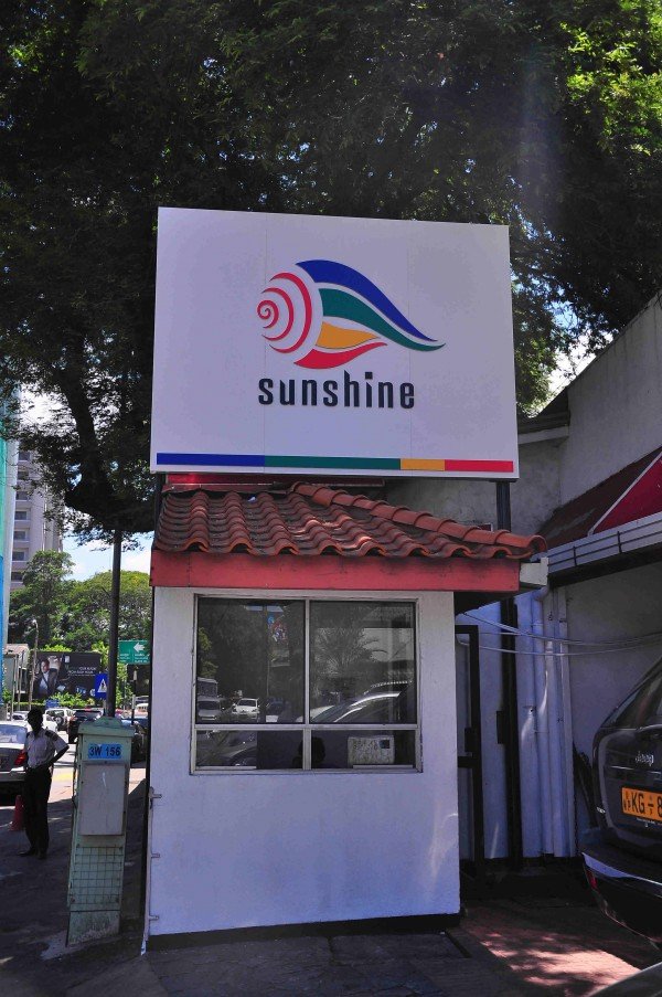 Signboard with the word 'Sunshine' and a colorful logo of a stylized sun with rainbow-colored waves, mounted above a small building with a red tiled roof.