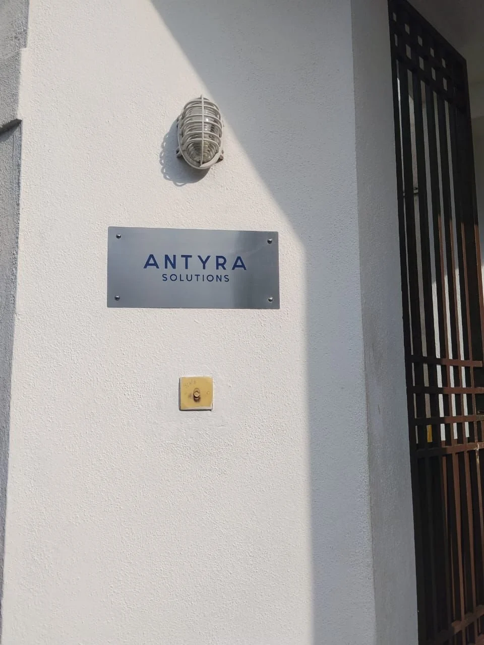 Exterior wall with a sign reading 'ANTYRA SOLUTIONS', an outdoor light fixture, and a yellow doorbell or intercom on a white wall.