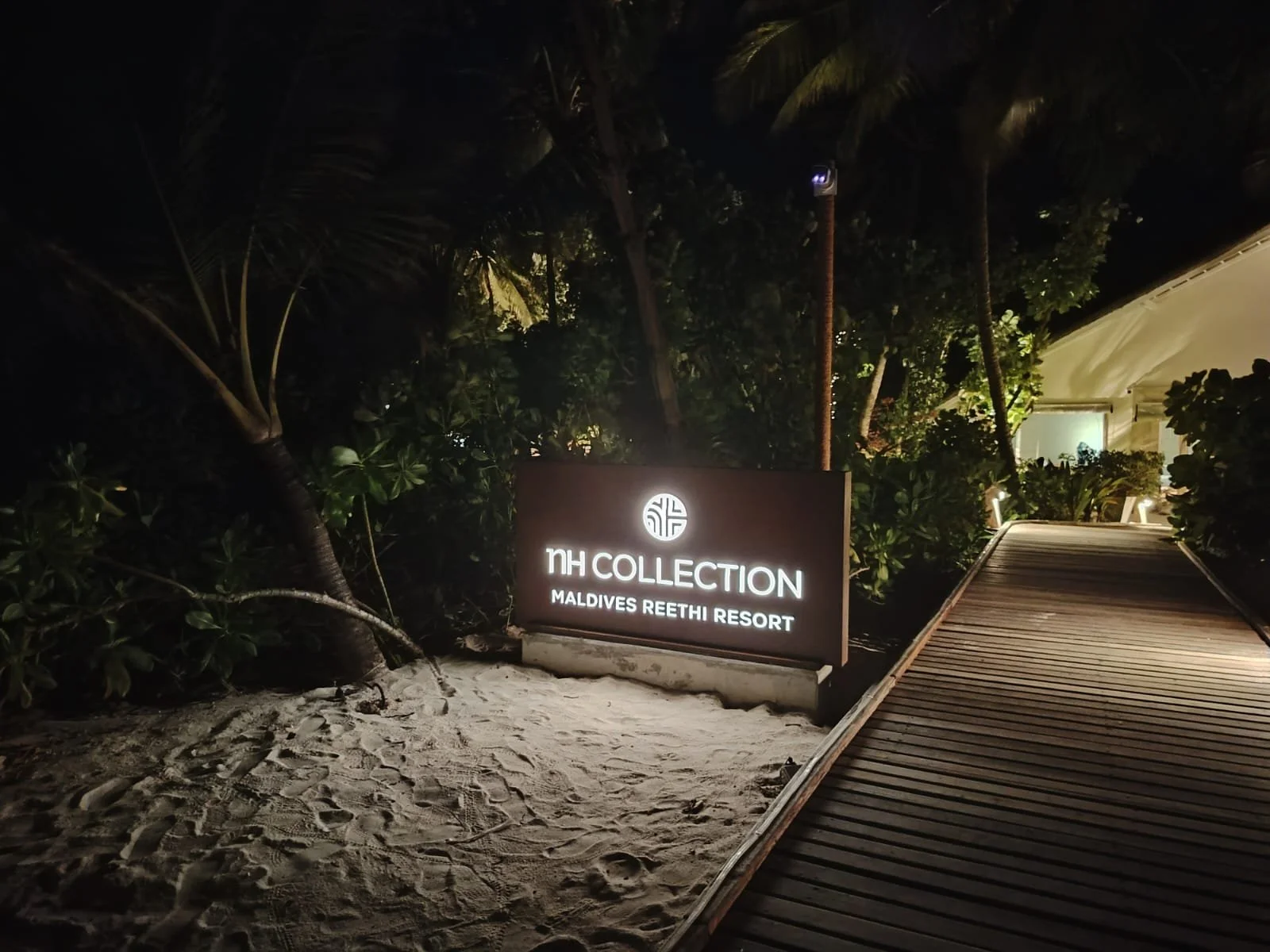 Nighttime view of the entrance sign for NH Collection Maldives Reethi Resort, illuminated, surrounded by palm trees, with a wooden walkway leading to the resort.