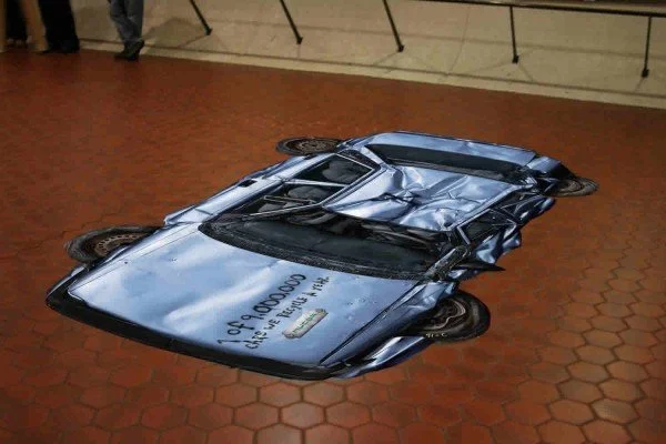 A crumpled silver sports car on display in an indoor setting.