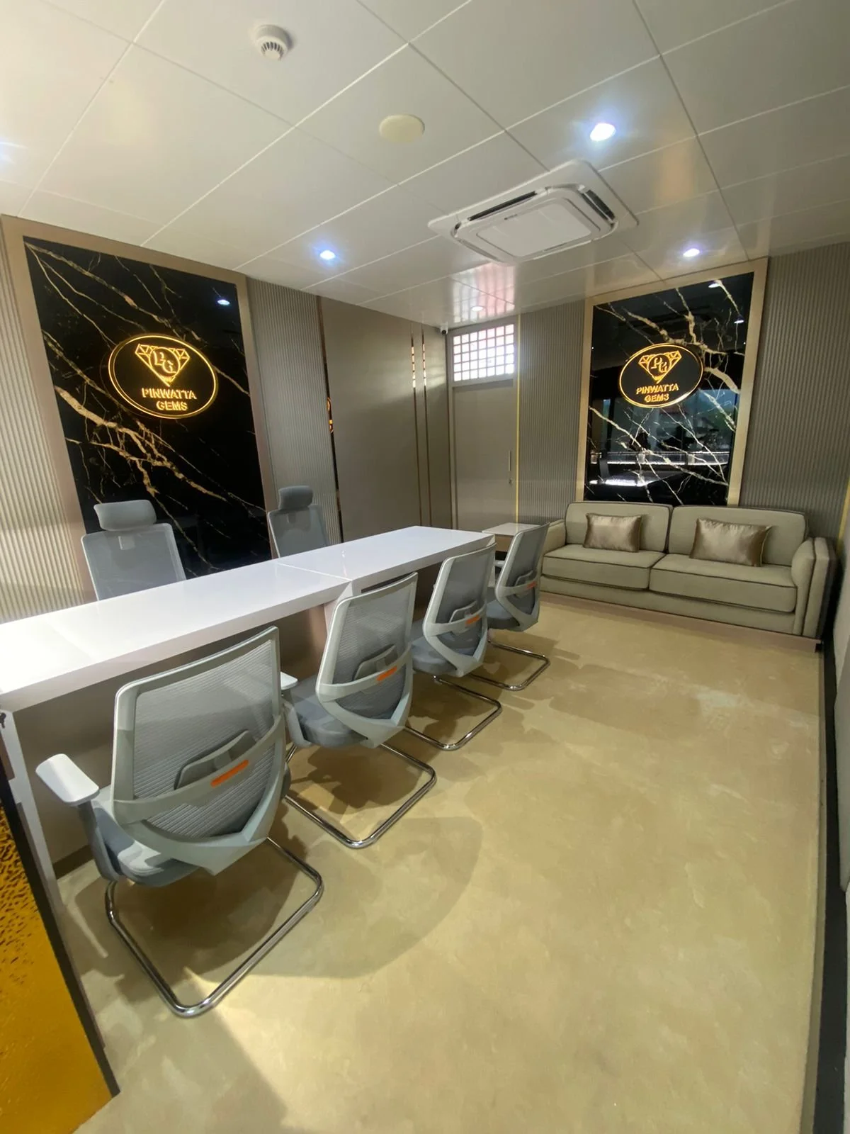 Modern office meeting room with a long white table, six gray swivel chairs, a beige sofa with three pillows, black marble accent walls with gold accents and illuminated signage reading 'PYNWATTA GEMS' with a diamond logo, beige marble flooring, ceili
