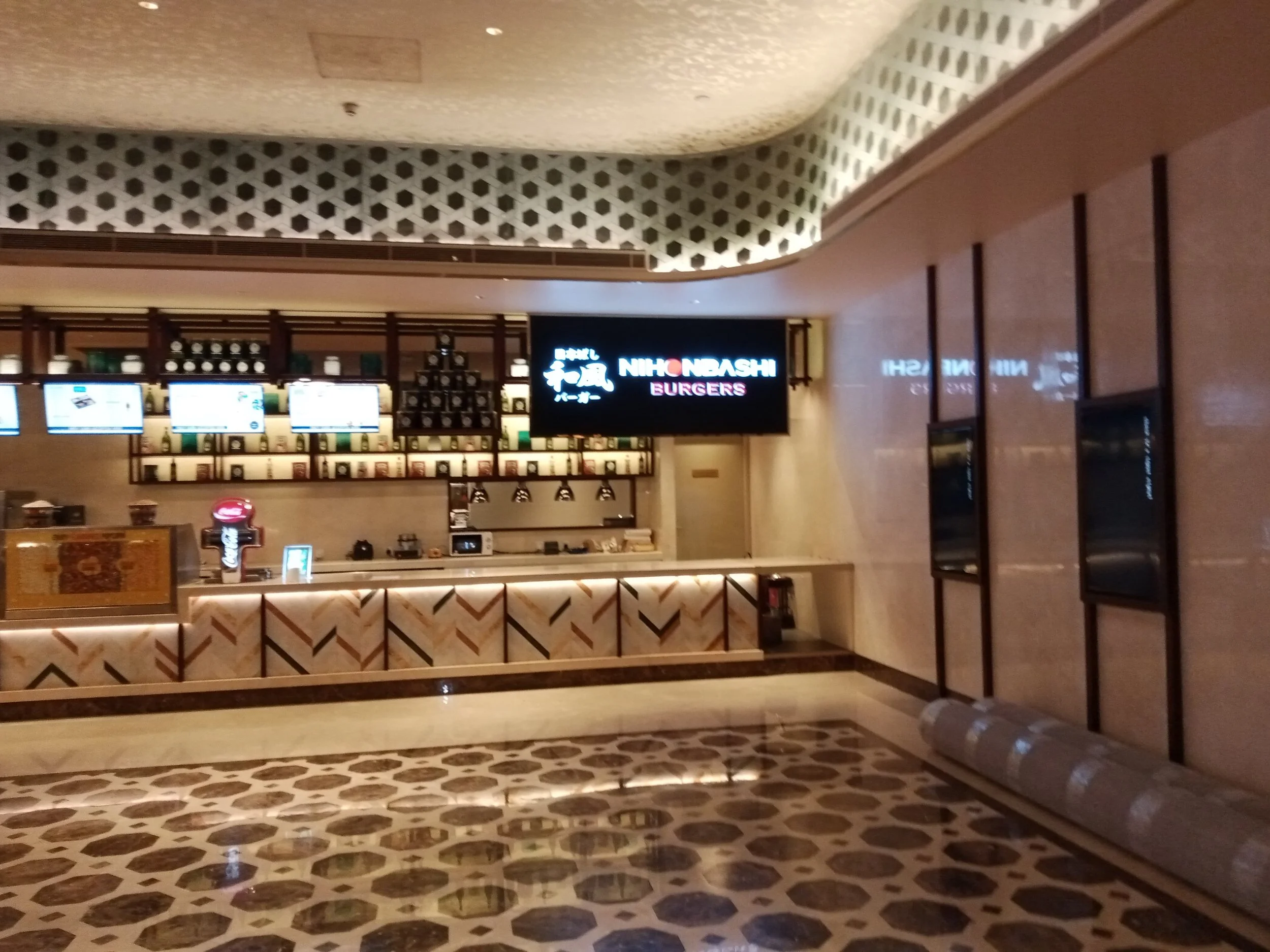 A lobby area featuring a wooden counter with a pattern of diagonal lines, a large digital menu board, and a neon sign reading 'Nihonbashi Burgers'. The ceiling has a decorative lattice pattern with built-in lighting, and the floor has a geometric til