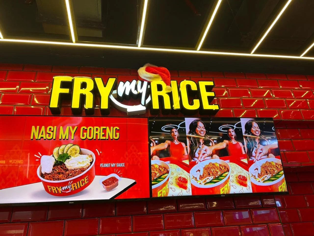 Signboard for Fry My Rice with a Santa hat on the text, showing a red background with tiled wall and attached digital menu screens featuring images of rice dishes and a woman in red.