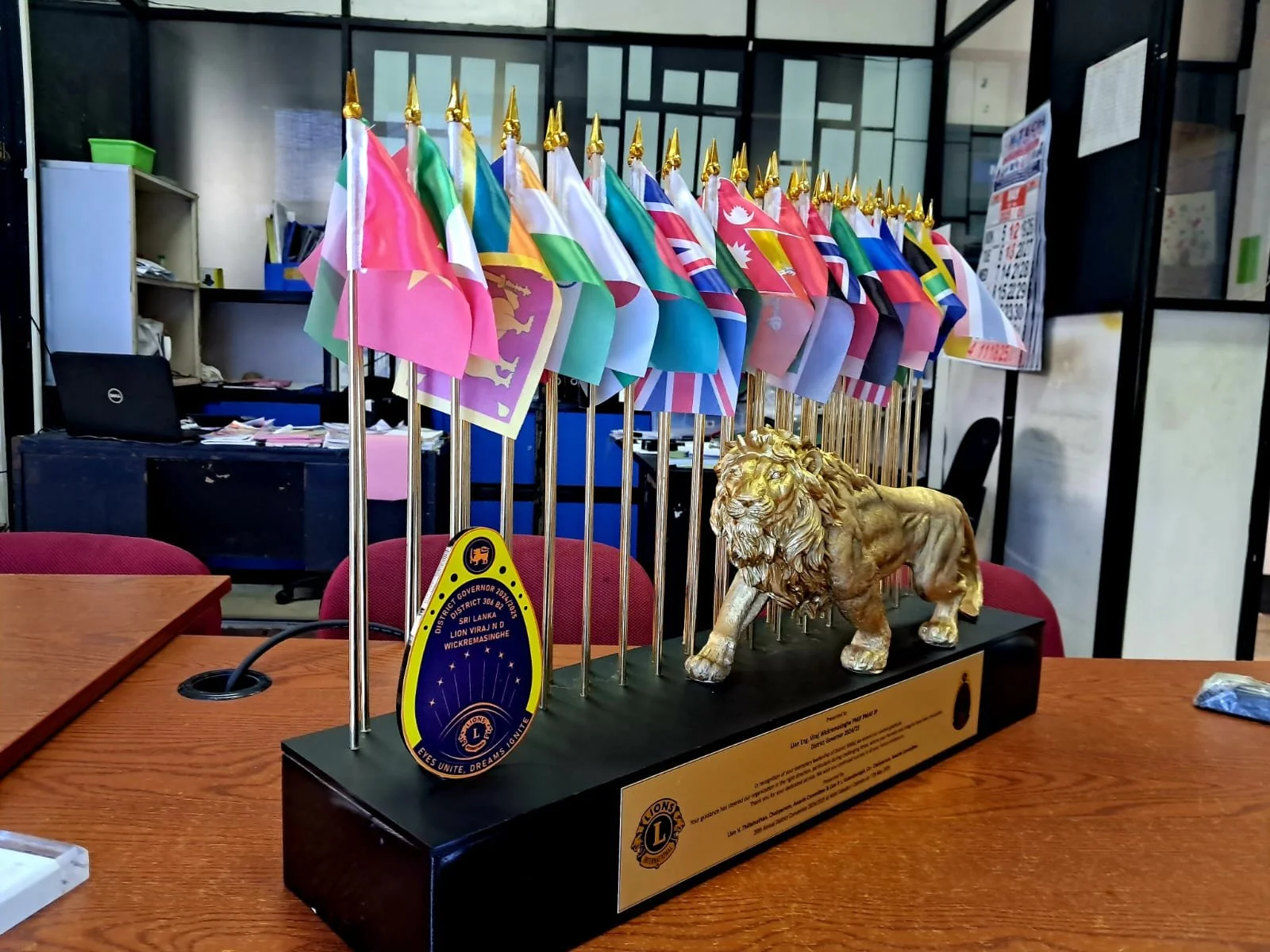 A Lions Club trophy with multiple small international flags on gold poles, and a gold lion figure, displayed on a wooden table in an office setting.
