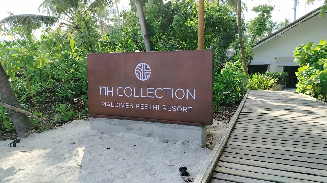 Sign for NH Collection Maldives Reethi Resort next to a wooden pathway surrounded by lush greenery and palm trees.