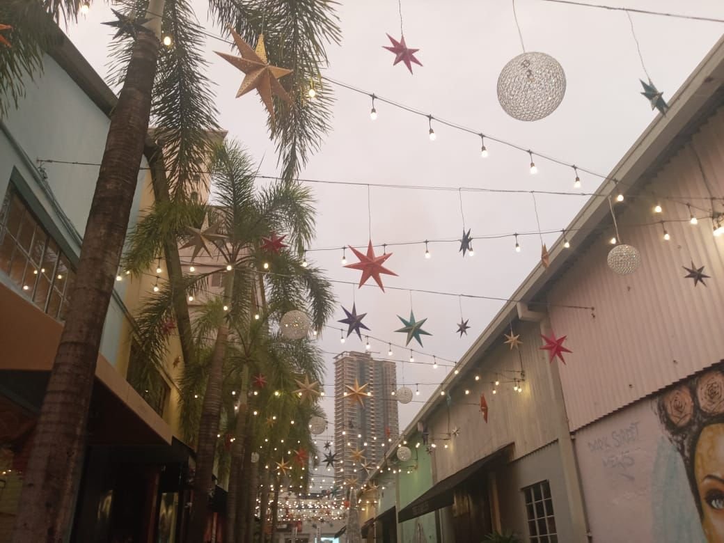 Outdoor alleyway decorated with hanging string lights, colorful star and spherical paper lanterns, and palm trees, with buildings visible in the background.