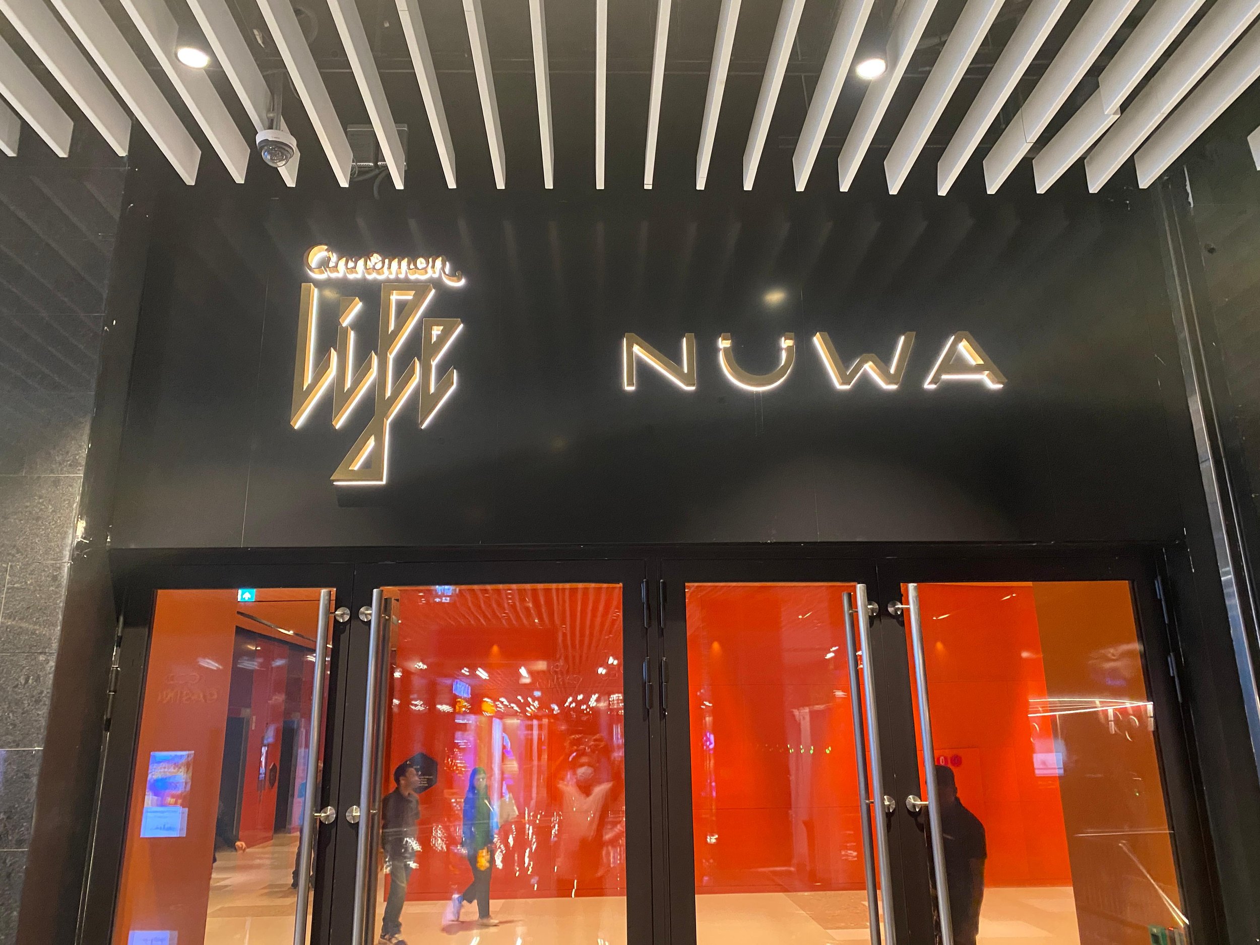 Entrance to a store called 'Nuwa' with glass doors and a black and gold logo sign above, inside the store red lighting and people walking around.