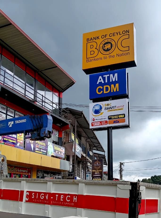 Signage indicating a Bank of Ceylon ATM and CDM in a shopping area with multiple commercial signs and buildings under cloudy skies.
