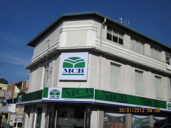 A corner building with MCB bank signage and branding, featuring a large MCB sign above the entrance and smaller sign with the bank's logo on the corner, under a clear blue sky.